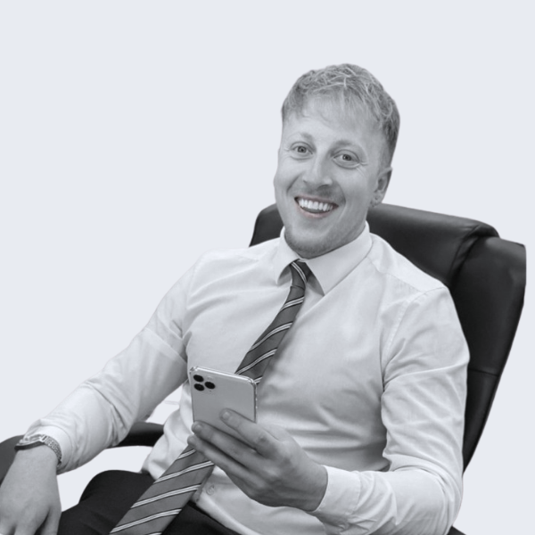 Photo shows a man with short blonde hair hairstyle, in a shirt and tie, leaning back in an office chair, holding a mobile phone in one hand and smiling at the camera. Representing a recruitment agency near you, in Cirencester.