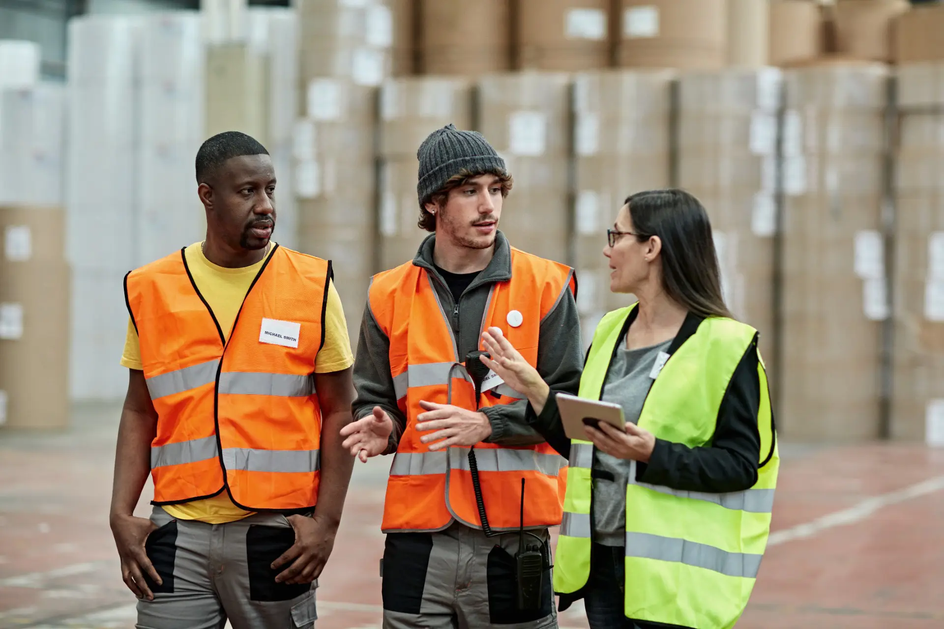 two men in orange high-visibility vests walking alongside a woman in a yellow high-visibility vest through a warehouse while talking