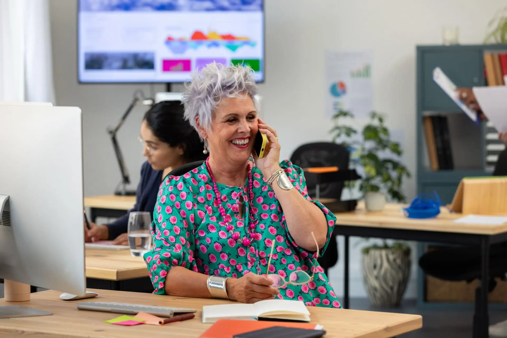 A woman with short, textured grey hair, wearing a colourful top and smiling whilst talking into her mobile, sits at her desk in an open office