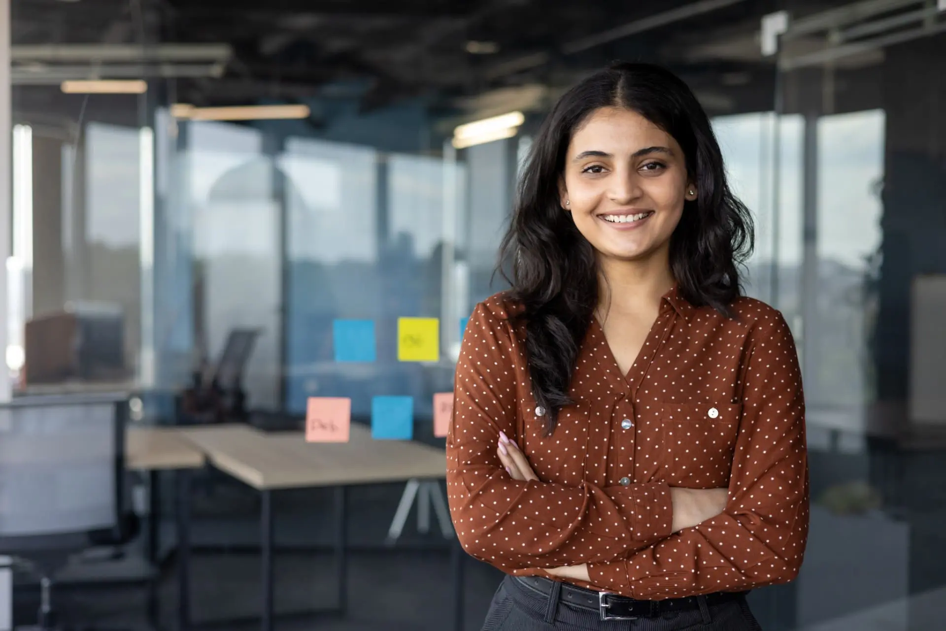 A woman in her 20s stands facing the camera, arms folded and smiling. She is in an open-plan office.