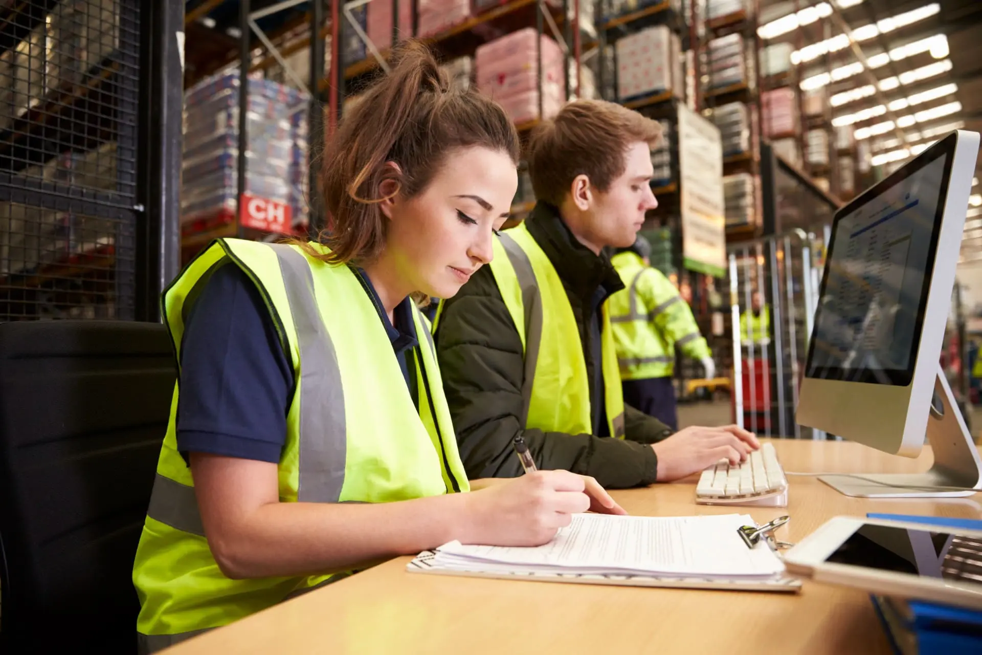 Image shows a woman and a man in high viz and casual clothing working in front of a pc at a desk inside a warehouse.