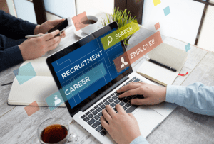 Hands typing at a laptop, words coming off the laptop screen which read recruitment, career, search, employee. Hands seen in background looking at a phone screen. 