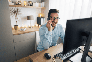 Brunette man in glasses and a light blue shirt, and smiling as he takes a call on a mobile phone. Working at a computer in a living room.