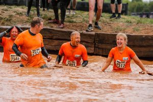 Men and women, in sports t-shirts and runner numbers laughing and smiling in muddy pool. 