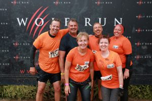 Group of 6 men and women in orange sports tops grouped for a photo. The background says Wolf Run and the runners are muddy. 